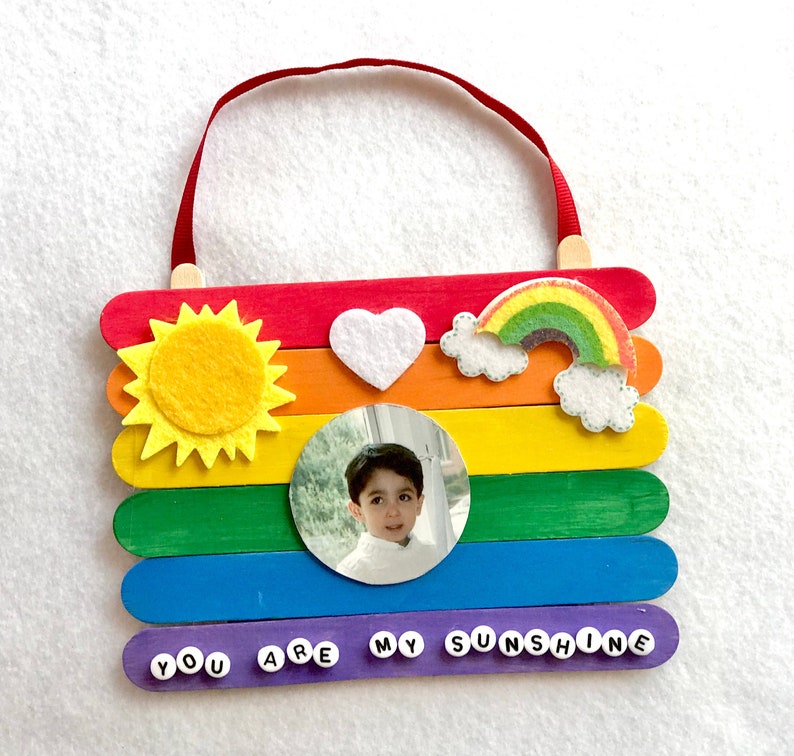 May include: A rainbow-colored popsicle stick craft with a photo of a child, a felt sun, a felt heart, and a felt rainbow. The bottom row of popsicle sticks has the words "You Are My Sunshine" spelled out with letter beads.