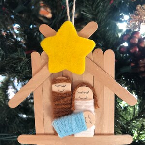 May include: A handmade Christmas bauble depicting the nativity scene. The bauble is made from craft sticks and features a yellow felt star, a brown yarn-wrapped figure of Joseph, a white yarn-wrapped figure of Mary, and a blue yarn-wrapped baby Jesus.