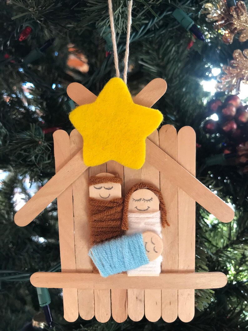 May include: A handmade Christmas bauble depicting the nativity scene. The bauble is made from craft sticks and features a yellow felt star, a brown yarn-wrapped figure of Joseph, a white yarn-wrapped figure of Mary, and a blue yarn-wrapped baby Jesus.