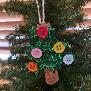 Pu&ograve; includere: Un ornamento natalizio fatto a mano a forma di albero. L'albero &egrave; realizzato con orpelli verdi e decorato con bottoni colorati in rosso, rosa, giallo, arancione e azzurro. Il tronco &egrave; un pezzo di cannella, appeso a uno spago bianco.