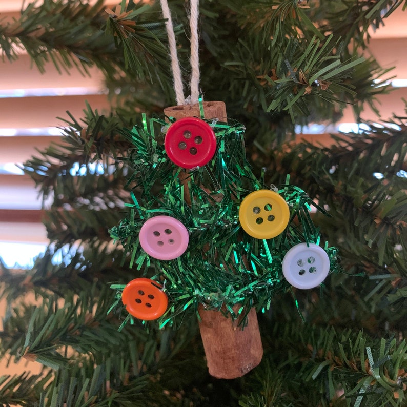 Pu&ograve; includere: Un ornamento natalizio fatto a mano a forma di albero. L'albero &egrave; realizzato con orpelli verdi e decorato con bottoni colorati in rosso, rosa, giallo, arancione e azzurro. Il tronco &egrave; un pezzo di cannella, appeso a uno spago bianco.
