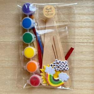 May include: A craft kit for making a rainbow unicorn with paint, wooden sticks, and felt pieces. The kit includes six small bottles of paint in red, orange, yellow, green, blue, and purple. The felt pieces include a rainbow, two clouds, and a sun. The kit also includes wooden sticks, a paintbrush, and letter beads that spell out "I love unicorns".