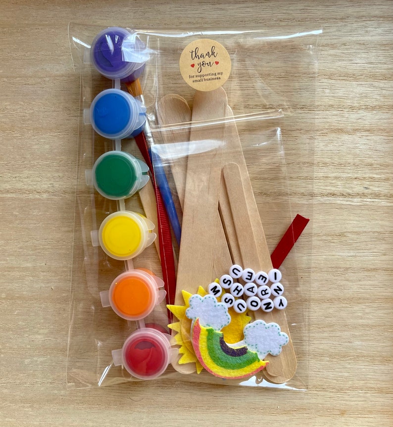 May include: A craft kit for making a rainbow unicorn with paint, wooden sticks, and felt pieces. The kit includes six small bottles of paint in red, orange, yellow, green, blue, and purple. The felt pieces include a rainbow, two clouds, and a sun. The kit also includes wooden sticks, a paintbrush, and letter beads that spell out "I love unicorns".