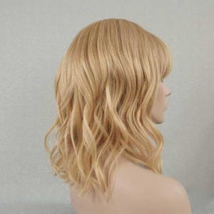 May include: A shoulder-length wig featuring wavy, light blonde hair with subtle golden highlights. The hair is styled with soft, loose waves that fall around the shoulders, and it includes a fringe that frames the forehead area in a natural manner.