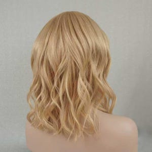 May include: A shoulder-length wig featuring wavy, light blonde hair with subtle golden highlights. The hair is styled with a soft, natural texture and a center part, falling in loose, flowing waves that frame the head and shoulders evenly.