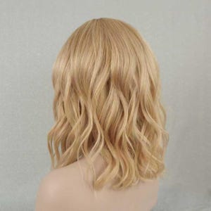 May include: A shoulder-length wig featuring wavy, light blonde hair with subtle golden highlights. The hair is styled with a natural-looking part and soft, loose waves that cascade down to the shoulders, creating a voluminous and textured appearance.