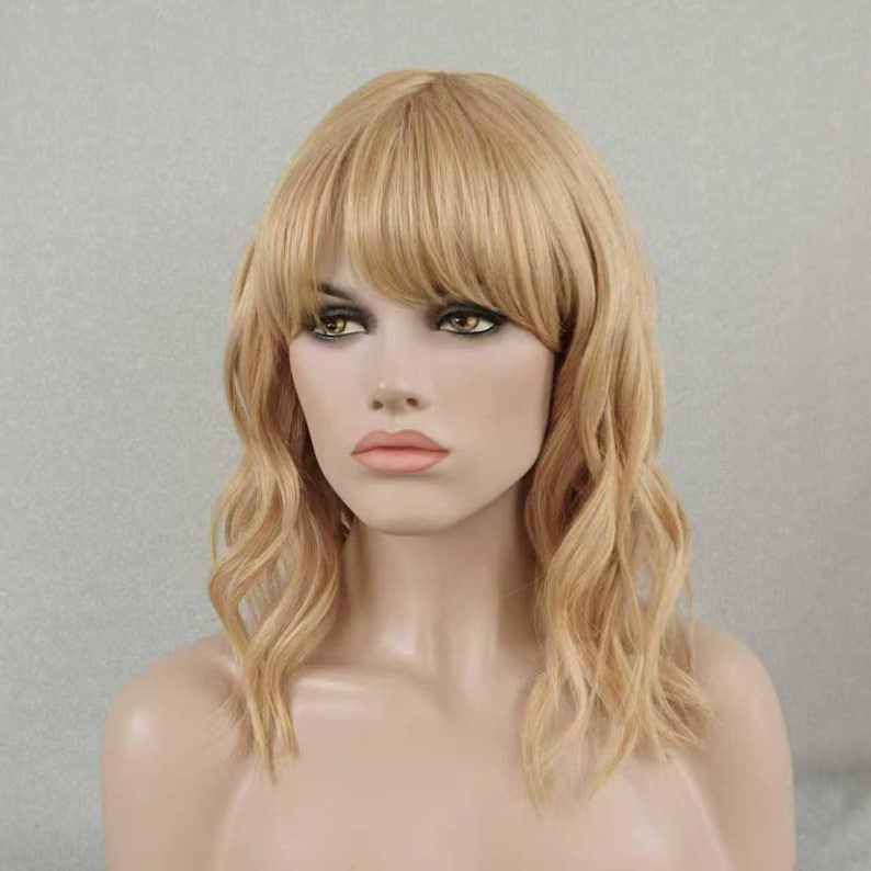 May include: A shoulder-length blonde wig featuring soft waves and straight bangs. The hair is a light golden shade with subtle highlights, styled to frame the face with a layered cut that falls just past the shoulders in a natural, wavy texture.