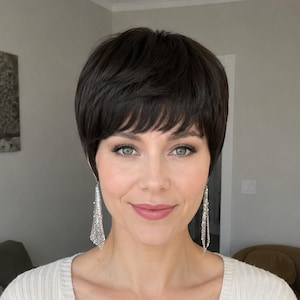 Peut inclure: Une femme aux cheveux courts et brun foncé avec une frange balayée sur le côté. Elle porte des boucles d'oreilles pendantes argentées. La femme sourit et regarde la caméra. L'arrière-plan est gris neutre.