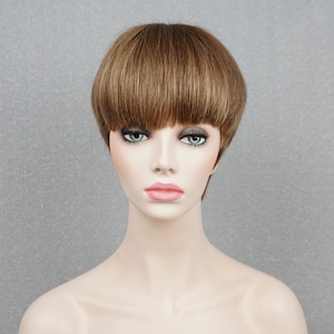 May include: A mannequin head displaying a short, layered wig with a blunt fringe. The wig features a blend of light brown and golden highlights, creating a natural look. The hair is styled to frame the face, offering a modern and chic appearance.