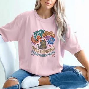 May include: Pale pink t-shirt featuring a cat illustration and the words "OVERTHINKING EVERYTHING AGAIN". The design includes thought bubbles with phrases such as "WHAT IF??" and "TOO MUCH?" in a playful font.