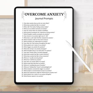 K&ouml;nnte beinhalten: Ein digitales Tablet zeigt "&Uuml;BERWINDE ANGST Journal-Fragen" mit einer Liste von Fragen. Das Tablet liegt auf einem Holztisch mit einem Stift, einer kleinen Pflanze und dekorativen Gegenst&auml;nden. Der Bildschirm ist schwarz mit wei&szlig;em Text.