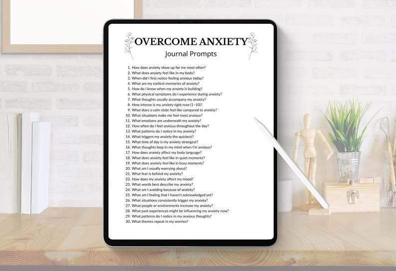 K&ouml;nnte beinhalten: Ein digitales Tablet zeigt "&Uuml;BERWINDE ANGST Journal-Fragen" mit einer Liste von Fragen. Das Tablet liegt auf einem Holztisch mit einem Stift, einer kleinen Pflanze und dekorativen Gegenst&auml;nden. Der Bildschirm ist schwarz mit wei&szlig;em Text.