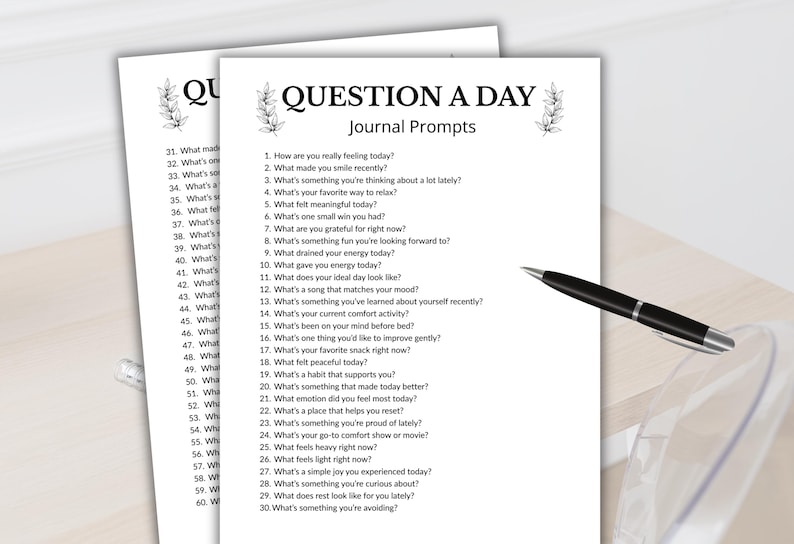 Może przedstawiać: Dwie białe strony z tekstem "QUESTION A DAY Journal Prompts" i numerowanymi pytaniami. Czarny długopis ze srebrną końc&oacute;wką spoczywa na stronach. Strony znajdują się na jasnej drewnianej powierzchni.