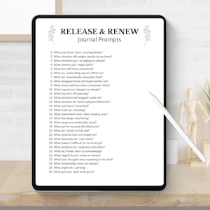 May include: A digital tablet displays "RELEASE & RENEW Journal Prompts" with a list of questions. The tablet is black with a white screen, and a white stylus rests nearby. The scene includes a wooden desk, a small plant, and decorative items.
