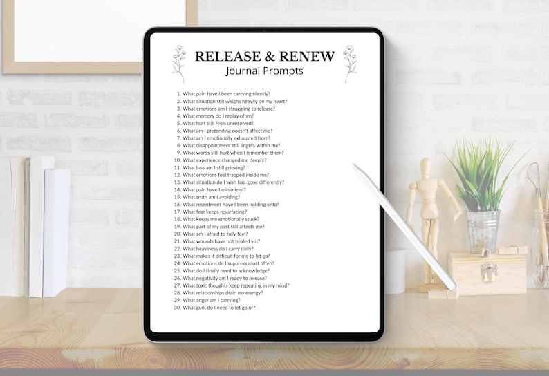 May include: A digital tablet displays "RELEASE & RENEW Journal Prompts" with a list of questions. The tablet is black with a white screen, and a white stylus rests nearby. The scene includes a wooden desk, a small plant, and decorative items.