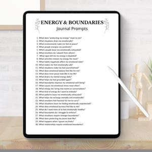 May include: A digital tablet displays "ENERGY & BOUNDARIES Journal Prompts" with a list of questions about emotional well-being. The tablet is black with a white stylus, set on a wooden desk with decorative items.