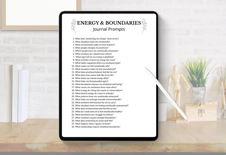 May include: A digital tablet displays "ENERGY & BOUNDARIES Journal Prompts" with a list of questions about emotional well-being. The tablet is black with a white stylus, set on a wooden desk with decorative items.