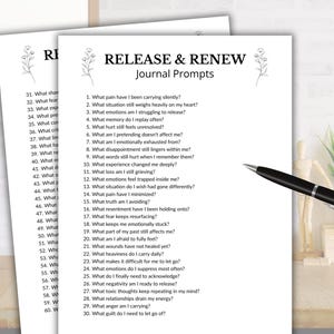 May include: Two white sheets of paper with journal prompts titled "RELEASE & RENEW." A black pen rests on the right side. The prompts are designed to encourage self-reflection and emotional processing.