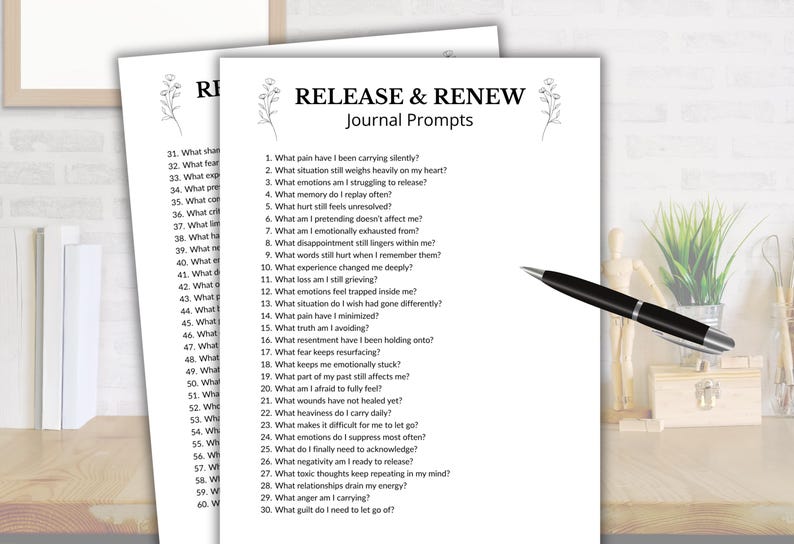 May include: Two white sheets of paper with journal prompts titled "RELEASE & RENEW." A black pen rests on the right side. The prompts are designed to encourage self-reflection and emotional processing.