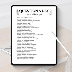 Może przedstawiać: Tablet cyfrowy wyświetla stronę dziennika z tekstem "QUESTION A DAY Journal Prompts". Pytania są ponumerowane i obejmują pytania dotyczące uczuć, wdzięczności i codziennych doświadczeń. Pi&oacute;ro leży na stole.