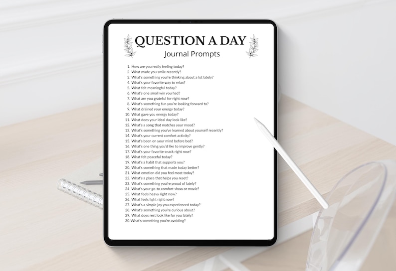 Może przedstawiać: Tablet cyfrowy wyświetla stronę dziennika z tekstem "QUESTION A DAY Journal Prompts". Pytania są ponumerowane i obejmują pytania dotyczące uczuć, wdzięczności i codziennych doświadczeń. Pi&oacute;ro leży na stole.