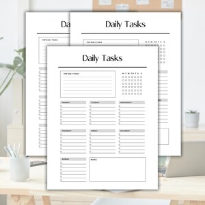 May include: Several white Daily Tasks planner sheets with a minimalist design. Each sheet features sections for top daily tasks, a weekly calendar, and daily task lists. The sheets are on a wooden desk with a plant and a laptop.