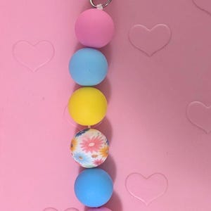 May include: A beaded keychain with a silver clasp. The keychain features a series of colorful, round beads in pink, blue, yellow, and a floral pattern. The background is a light pink surface with heart shapes.