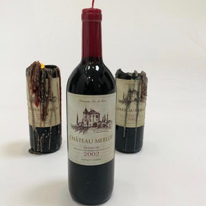 Puede incluir: Tres velas de botella de vino de novedad, dos están derretidas y una está llena. La vela llena está etiquetada "Chateau Merlot Grand Cru 2002" y "Vela de novedad".