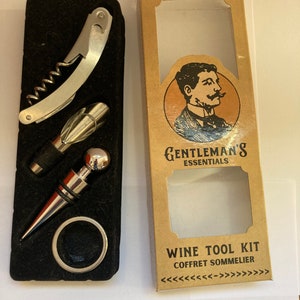May include: A wine tool kit with a corkscrew, bottle stopper, and aerator on a black base. The kit includes a silver bottle ring and is packaged in a cardboard box with the text "GENTLEMAN'S ESSENTIALS" and "WINE TOOL KIT".