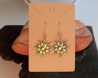 May include: A pair of gold-colored sunburst earrings with a hook closure. The earrings are displayed on a tan earring card, set against a natural stone surface. The sun design features a central circle with radiating rays.