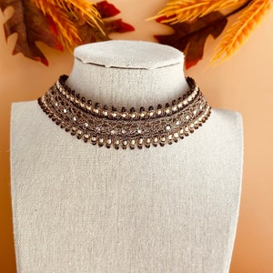 May include: A brown and gold choker necklace with a beaded design. The necklace is on a white mannequin.