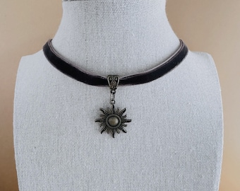 May include: A black velvet choker necklace with a gold-toned sun pendant.