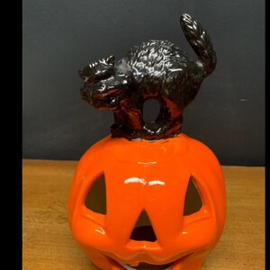 Può includere: Una zucca in ceramica con una statuetta di gatto nero in cima. La zucca è arancione e ha un viso intagliato. Il gatto è in posizione accovacciata con la coda arrotolata attorno al suo corpo.