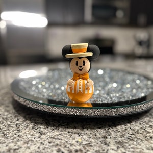 Puede incluir: Una pequeña figura naranja y negra de Mickey Mouse con un sombrero blanco y amarillo. La figura está de pie sobre una bandeja redonda negra y plateada.