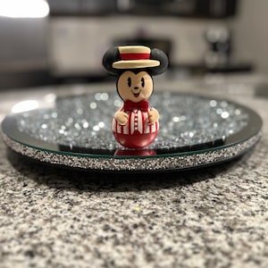 Puede incluir: Una figurita de Mickey Mouse a rayas rojas y blancas con un sombrero negro y una pajarita. La figurita está de pie sobre una bandeja negra y plateada con una superficie de espejo.