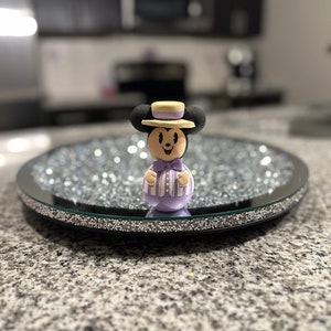 Puede incluir: Una figurita de Mickey Mouse a rayas moradas y blancas con un sombrero amarillo y una pajarita. La figurita está de pie sobre una bandeja redonda, negra y de espejo con un borde de purpurina plateada.