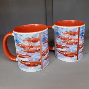 Op de afbeelding: Twee oranje en witte keramische mokken met een ontwerp van rode vliegtuigen die in formatie vliegen tegen een blauwe en witte achtergrond. De mokken hebben een rode handgreep.