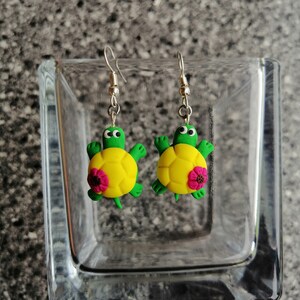 Op de afbeelding: Een paar hangende oorbellen met een groene en gele schildpad met een roze bloem op zijn schild. De oorbellen zijn gemaakt van polymer clay en hebben zilveren haakjes.