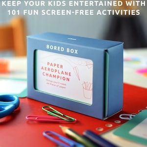 May include: A blue box with the text "BORED BOX" on the front. The box contains cards with activities for kids. One card is visible and says "PAPER AEROPLANE CHAMPION" with the text "Things you'll need: A piece of paper".