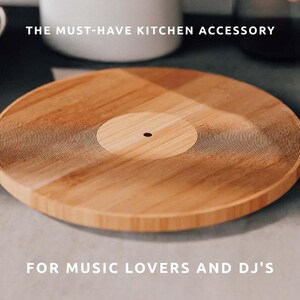 K&ouml;nnte beinhalten: Rundes Schneidebrett aus hellem Holz mit Vinyl-Schallplatten-Design. Das Brett hat konzentrische Rillen und einen dunklen Mittelpunkt. Der Text lautet "THE MUST-HAVE KITCHEN ACCESSORY" und "FOR MUSIC LOVERS AND DJ'S."