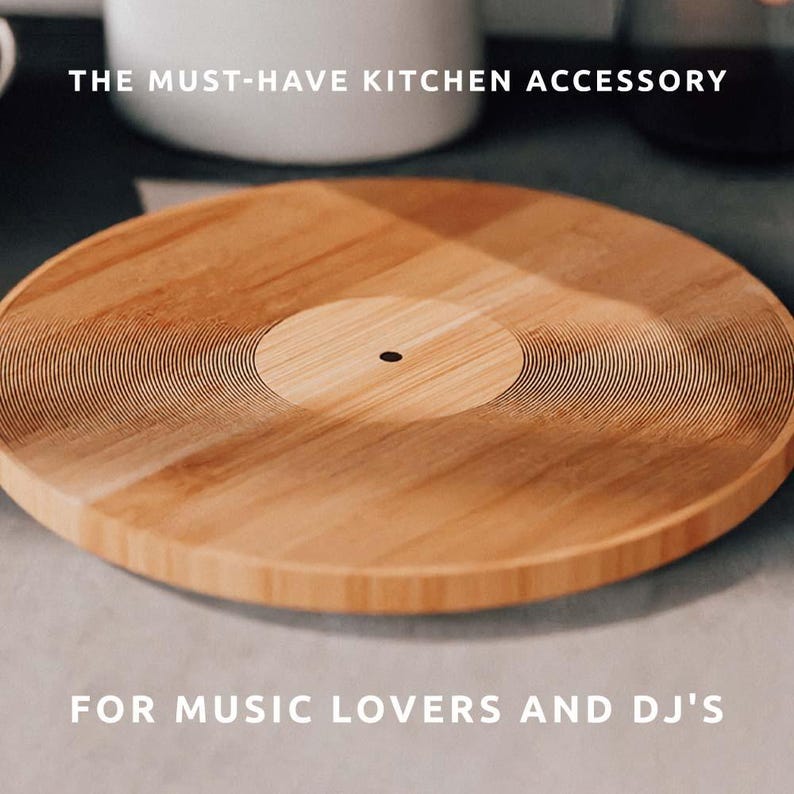 K&ouml;nnte beinhalten: Rundes Schneidebrett aus hellem Holz mit Vinyl-Schallplatten-Design. Das Brett hat konzentrische Rillen und einen dunklen Mittelpunkt. Der Text lautet "THE MUST-HAVE KITCHEN ACCESSORY" und "FOR MUSIC LOVERS AND DJ'S."