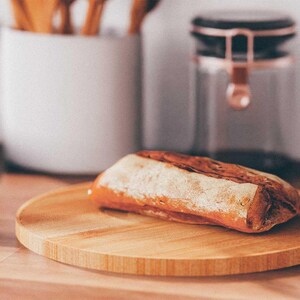 K&ouml;nnte beinhalten: Ein Laib knuspriges Brot liegt auf einem runden Holzschneidebrett. Das Brot hat eine goldbraune Kruste und eine leichte, mehlige Best&auml;ubung. Ein wei&szlig;er Keramik-Utensilienhalter und ein Glas mit kupferfarbenem Deckel befinden sich im Hintergrund.