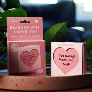 May include: A pink box with the text "Reasons Why I Love You" and a smaller pink box with a heart-shaped cutout and the text "You always make me laugh" inside. The boxes are on a pink and white surface.