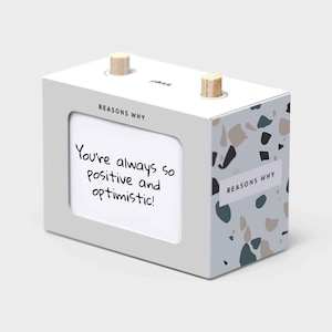 May include: A white box with a terrazzo pattern on the side. The box has a window that displays a card with the text "You're always so positive and optimistic!" The box has a wooden knob on each side. The text "Reasons Why" is printed on the front and side of the box.