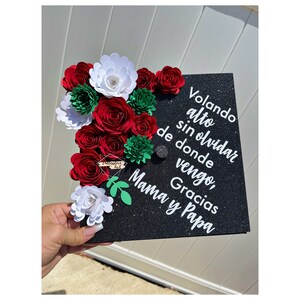 May include: Black graduation cap decorated with red and white paper flowers and green leaves. The cap has white text that reads "Volando alto sin olvidar de donde vengo, Gracias Mama y Papa".