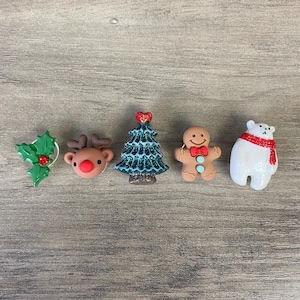 Puede incluir: Una colección de adornos navideños. Incluye una ramita de acebo, una cabeza de reno con nariz roja, un árbol de Navidad azul brillante, un hombre de jengibre con pajarita roja y un oso polar con bufanda roja.