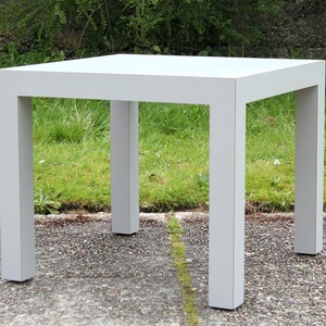 May include: A white square end table with a simple design. The table has four legs and a flat top.