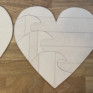 Puede incluir: Dos letreros de madera en forma de corazón sin terminar. Un corazón está en blanco, y el otro tiene un diseño de tres olas con líneas dibujadas en él.