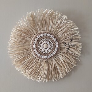 May include: Round wall decor featuring a central design of seashells on a crocheted tan circle, surrounded by natural-colored raffia. The piece has a bohemian aesthetic, with the text "Boho.ua" in the lower right corner.