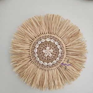 May include: A round, natural-toned woven wall hanging with a central design. The center features a star-shaped design made of shells, surrounded by a ring of shells and a woven border. The outer edge has a fringe.